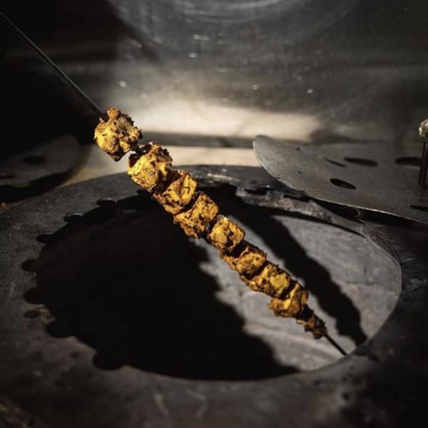 Indian paneer tikka skewers cooking inside a dark tandoor oven, with a metal lid nearby.
