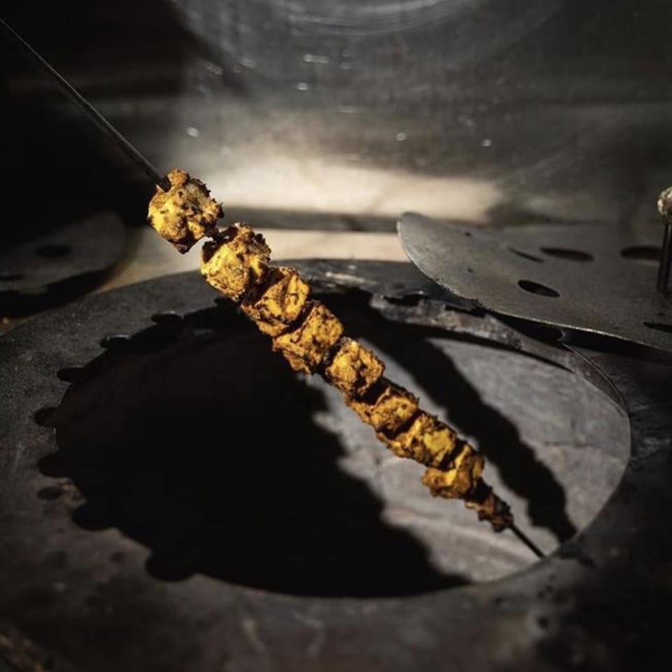 Indian paneer tikka skewers cooking inside a dark tandoor oven, with a metal lid nearby.