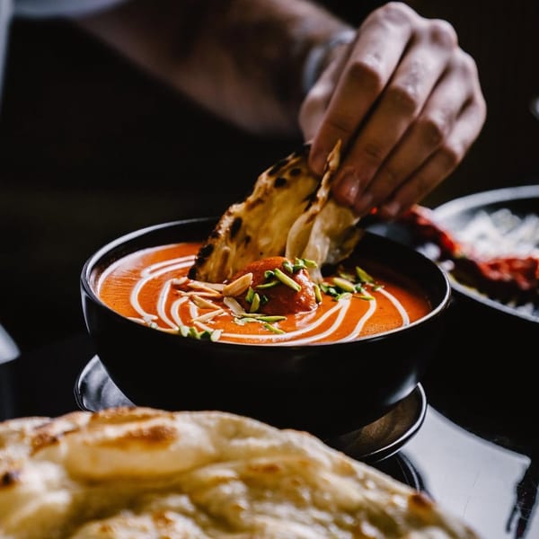Hand dipping naan bread into a bowl of vibrant orange Indian curry with cream swirls and nuts.