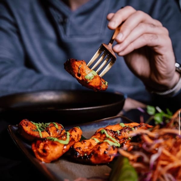 A hand uses a fork to lift a piece of Indian tandoori chicken with green sauce from a plate.