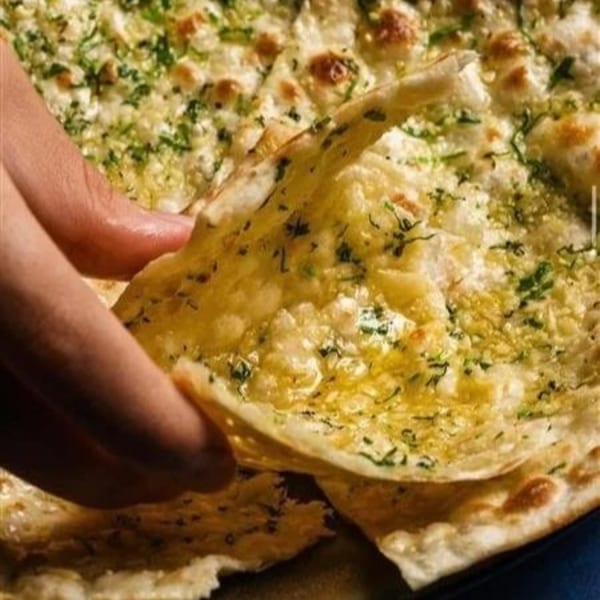 A hand pulls a piece of golden-brown garlic naan, glistening with butter and herbs, from a stack.