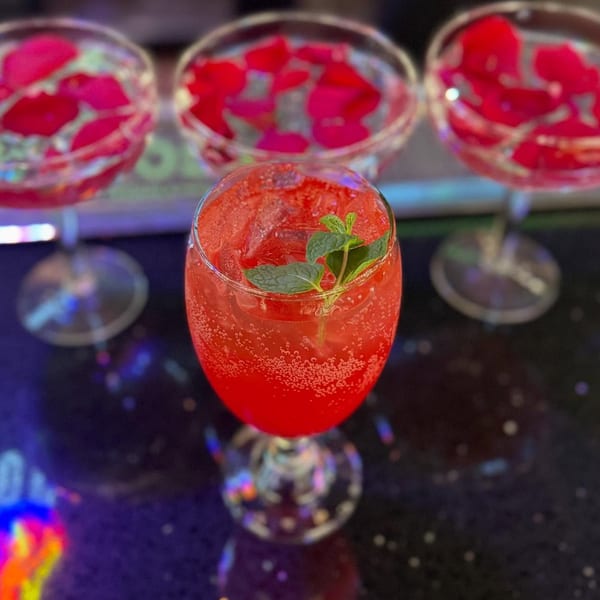 A vibrant red, bubbly cocktail with mint garnish, with two blurred glasses of rose petals in the background.