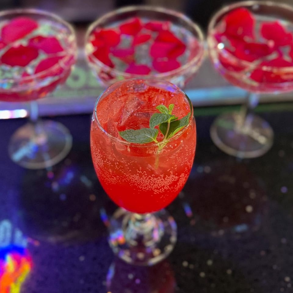 A vibrant red, bubbly cocktail with mint garnish, with two blurred glasses of rose petals in the background.
