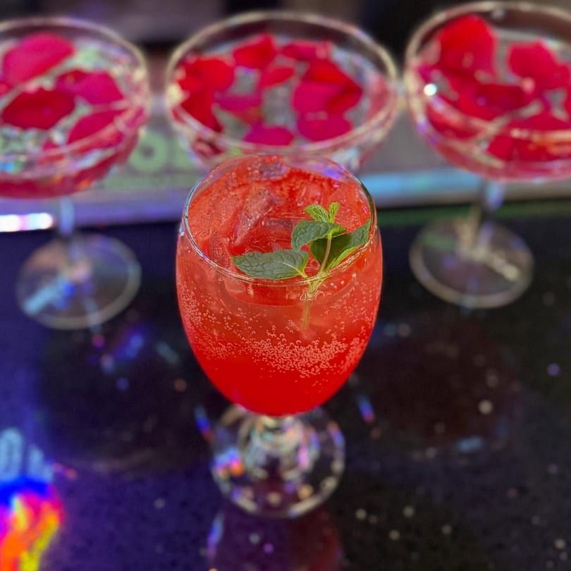 A vibrant red, bubbly cocktail with mint garnish, with two blurred glasses of rose petals in the background.