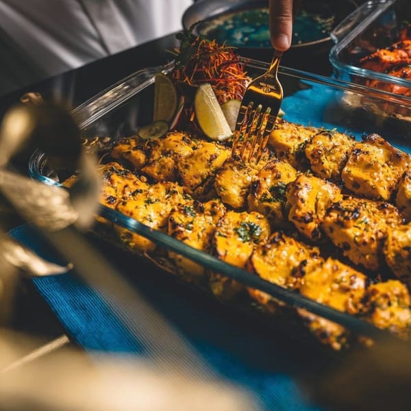 A golden fork piercing a piece of Indian chicken tikka in a glass dish with lime wedges.