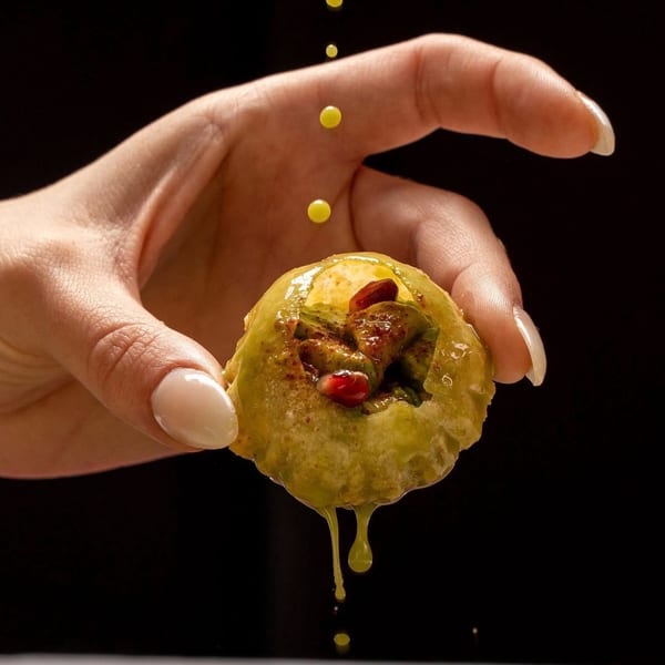 Hand holding a pani puri, filled with green chutney and pomegranate seeds, with yellow liquid dripping.