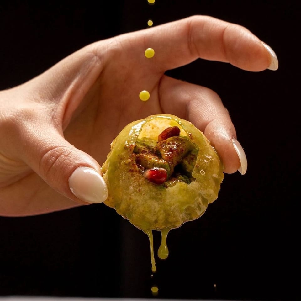 Hand holding a pani puri, filled with green chutney and pomegranate seeds, with yellow liquid dripping.