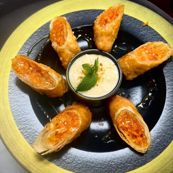 Six crispy Indian spring rolls with orange filling arranged around a bowl of creamy dip with mint.