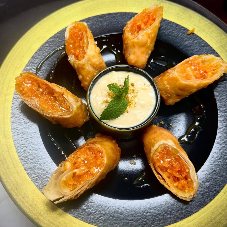 Six crispy Indian spring rolls with orange filling arranged around a bowl of creamy dip with mint.