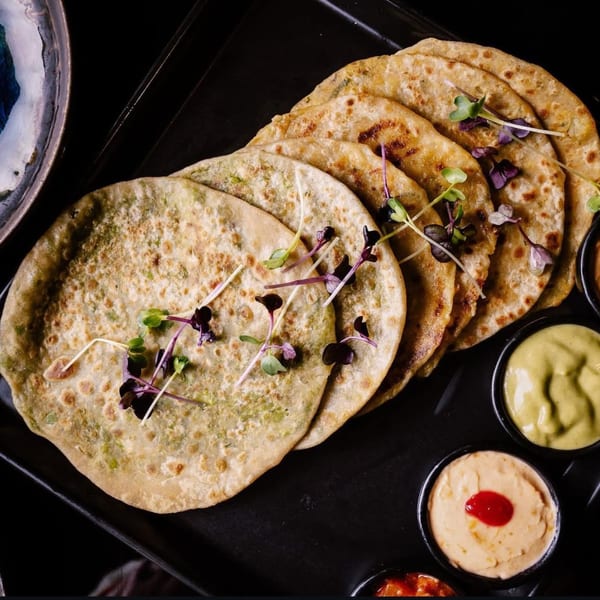 Indian parathas garnished with microgreens, served with three dipping sauces on a black tray.