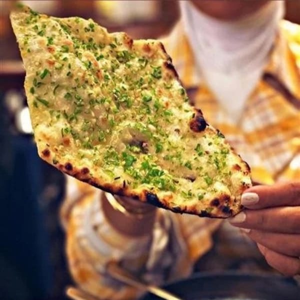 A person holding a piece of Indian garlic naan bread, charred and topped with green herbs.