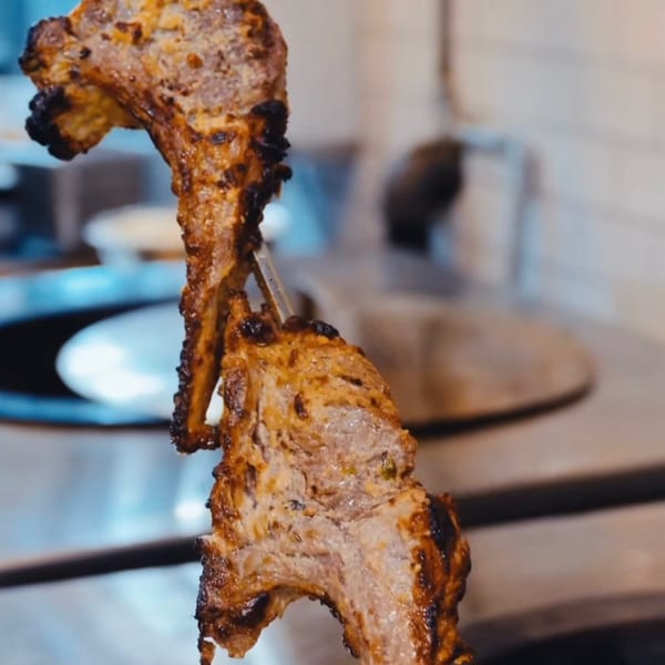 Two pieces of grilled Indian lamb chops on skewers, with a blurred kitchen background.