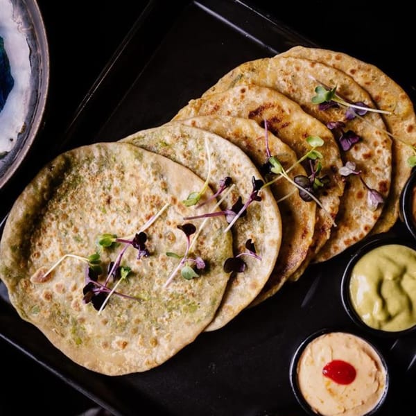 Indian parathas garnished with microgreens, served with two dipping sauces on a dark tray.