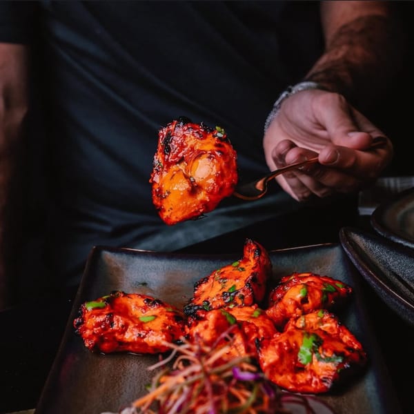 A hand uses a fork to lift a piece of vibrant red Indian chicken tikka from a dark plate.