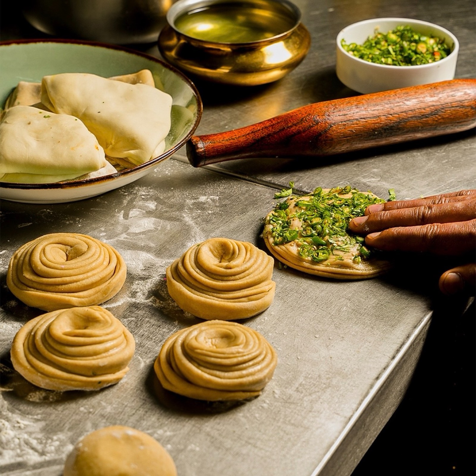 Unbaked spiral pastries on a floured counter beside filled dough, a rolling pin, chopped herbs, and bowls of ingredients.