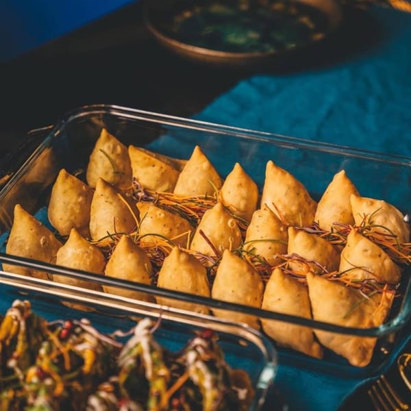 Golden-brown Indian samosas, garnished with herbs, arranged in a clear glass baking dish on a blue tablecloth.
