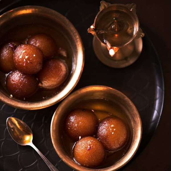 Two bowls of Gulab Jamun dessert with a spoon and a lit oil lamp on a dark tray.