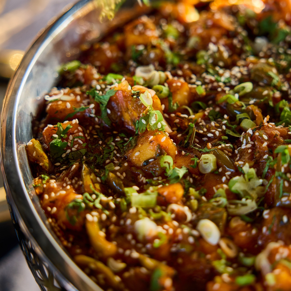 Close-up of a richly spiced saucy dish garnished with sesame seeds, scallions, and fresh cilantro in a metal bowl.