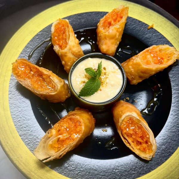 Six golden-fried Indian spring rolls with orange filling, drizzled with sauce, surrounding a creamy dip.